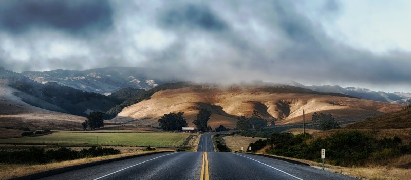 A lonely California road winding through the foothills while overhead clouds are building into a storm.